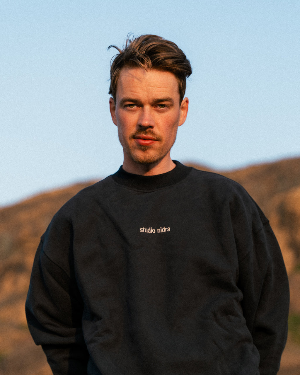 Man wearing The Crew Sweatshirt in black with Studio Nidra logo against mountainous background